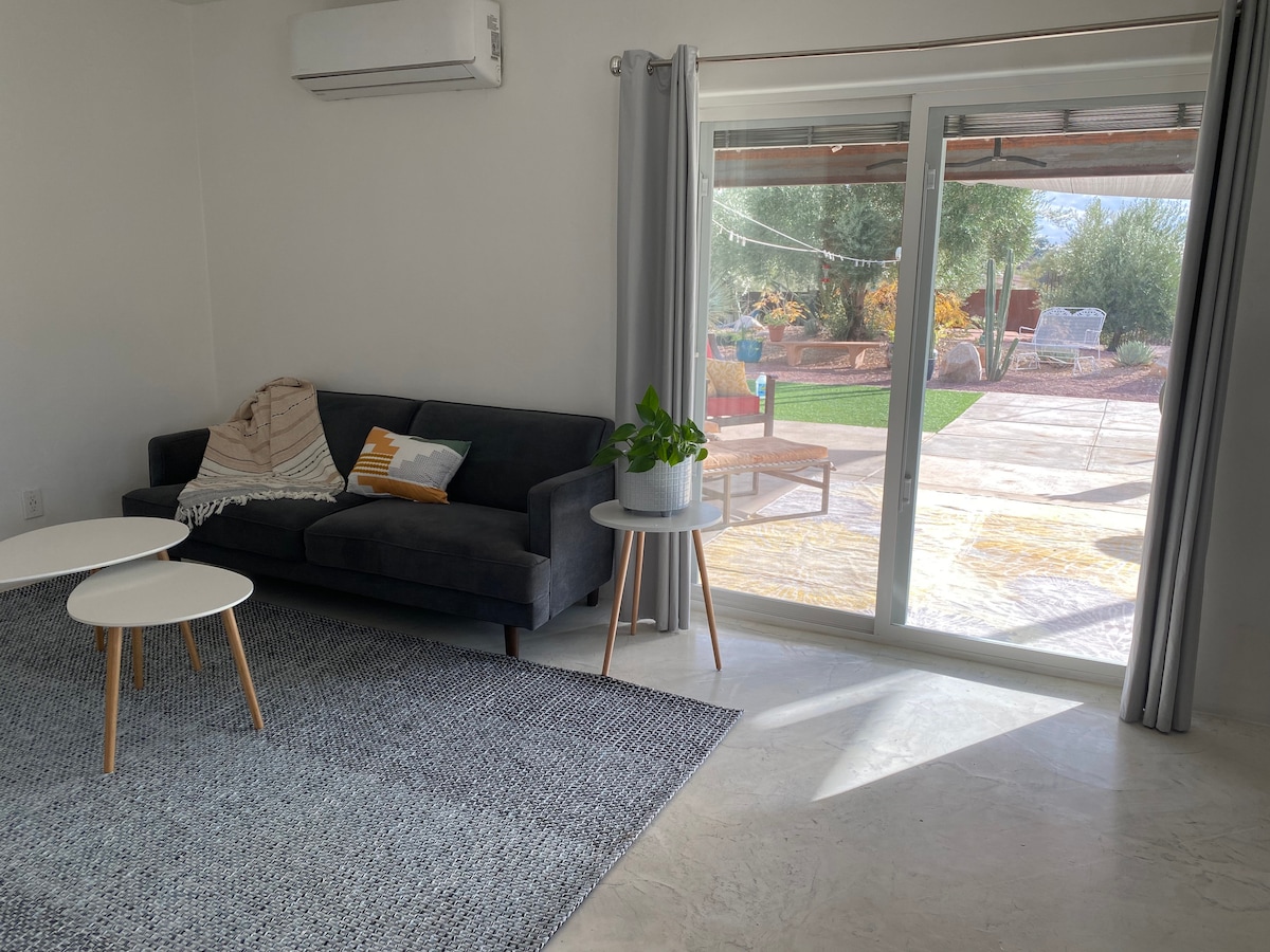 A cozy living area features a dark sofa with a light throw draped over one arm and a textured area rug beneath. Two small side tables are positioned beside the sofa. Sliding glass doors provide a view of the outdoor space, inviting natural light into the room.