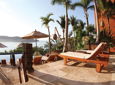 A sunlit terrace is adorned with lounge chairs and umbrellas, providing views of a tranquil water feature and lush palm trees. Stone flooring complements the natural elements, enhancing the inviting outdoor space designed for relaxation.