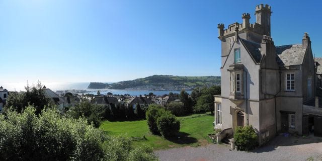 A grand tower section of Yannon Towers is visible against a clear blue sky. The coastal landscape stretches in the background, showcasing the sea and rolling hills. Lush greenery surrounds the property, enhancing its historical elegance.