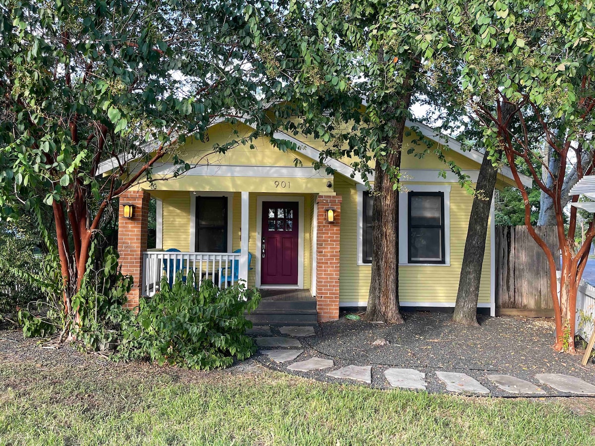 The front exterior of the bungalow is framed by lush trees and features a welcoming yellow facade. A red door is centered on the porch, flanked by white railings. A stone pathway leads to the entrance, while a fenced lawn provides an enclosed outdoor space.