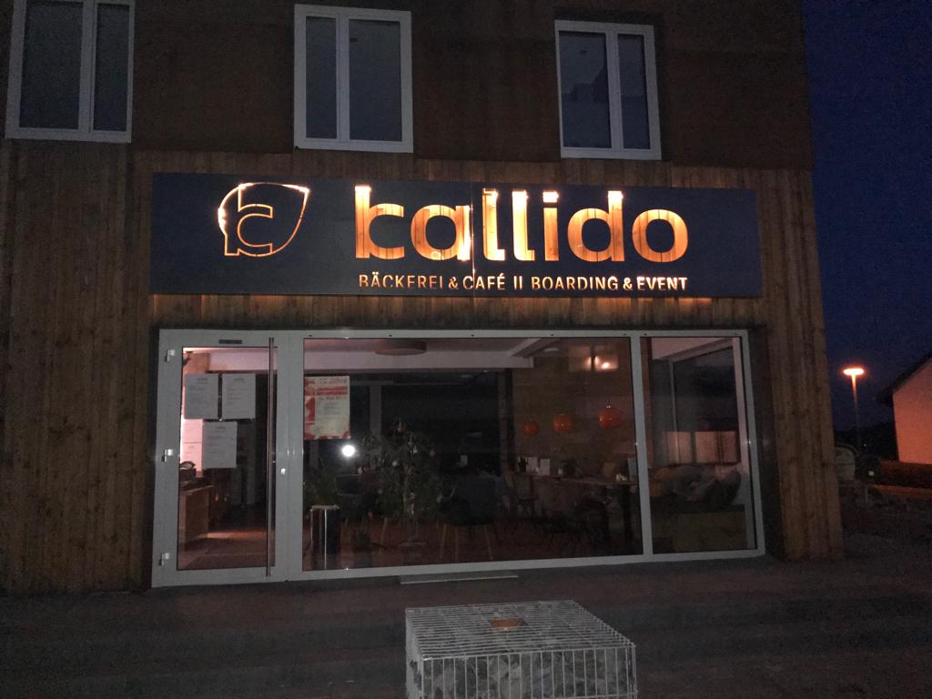 A modern building exterior is shown at dusk, featuring a well-lit sign displaying the name 'callido.' Large glass windows reveal the interior, where soft lighting creates a warm ambiance. The surrounding area is paved, providing accessibility, and an illuminated pathway leads to the entrance.