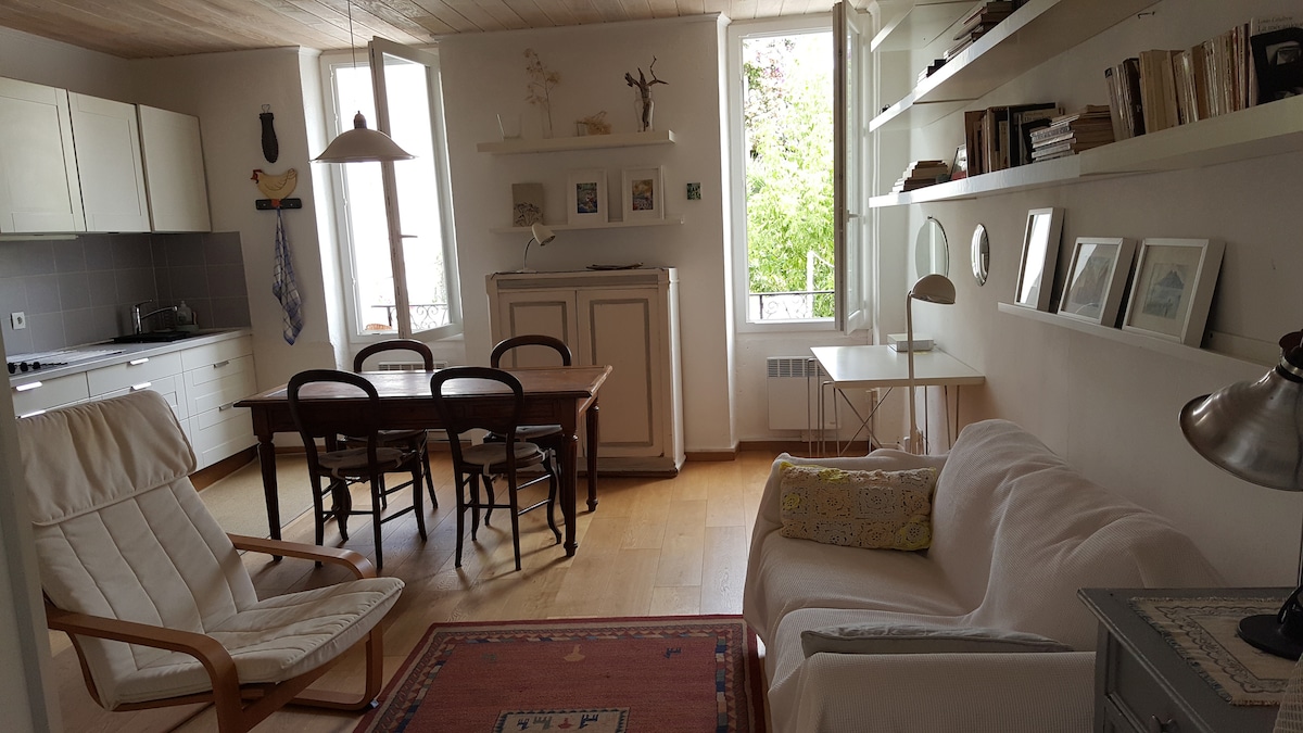 A bright living area features a cozy sofa and a comfortable armchair. A wooden dining table with black chairs is positioned nearby. Large windows invite natural light, while a shelving unit displays decorative items. Light-colored walls and wooden flooring create a welcoming space.