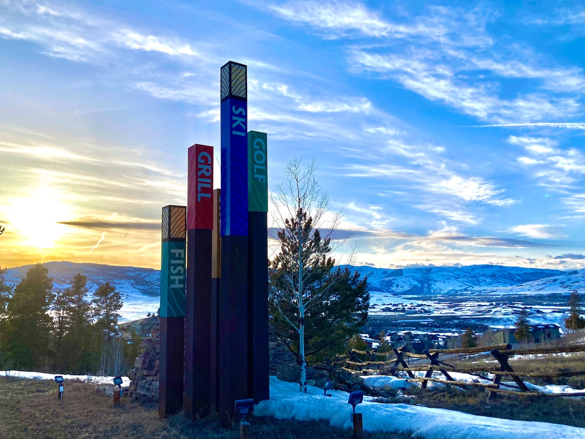 A row of colorful totem-like signs stands against a backdrop of a sunset sky, displaying the words 'Ski,' 'Grill,' 'Fish,' and 'Golf.' Snow covers the ground, and distant snow-capped mountains are visible, framed by trees and a wooden fence.