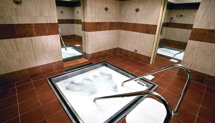 A spacious hot tub is positioned within a tiled room, featuring warm-toned walls and flooring. Handrails are installed for support, and the area is equipped with other amenities visible in the mirrors reflecting the soft lighting.