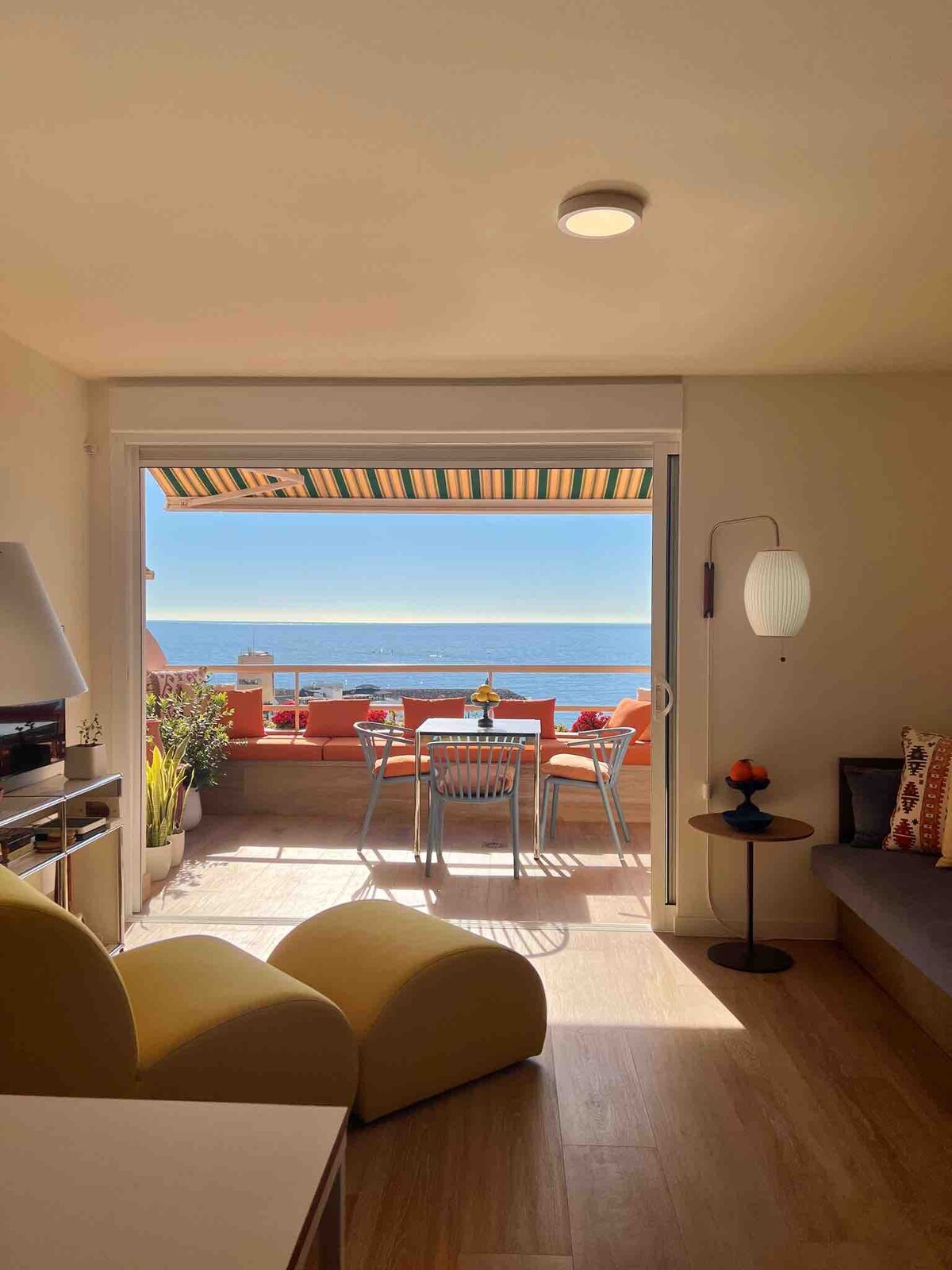 A living area features a view of the ocean through sliding glass doors. A small dining set is arranged on the terrace, with vibrant outdoor seating and decorative plants. Soft natural light illuminates the space, creating a warm, inviting environment.