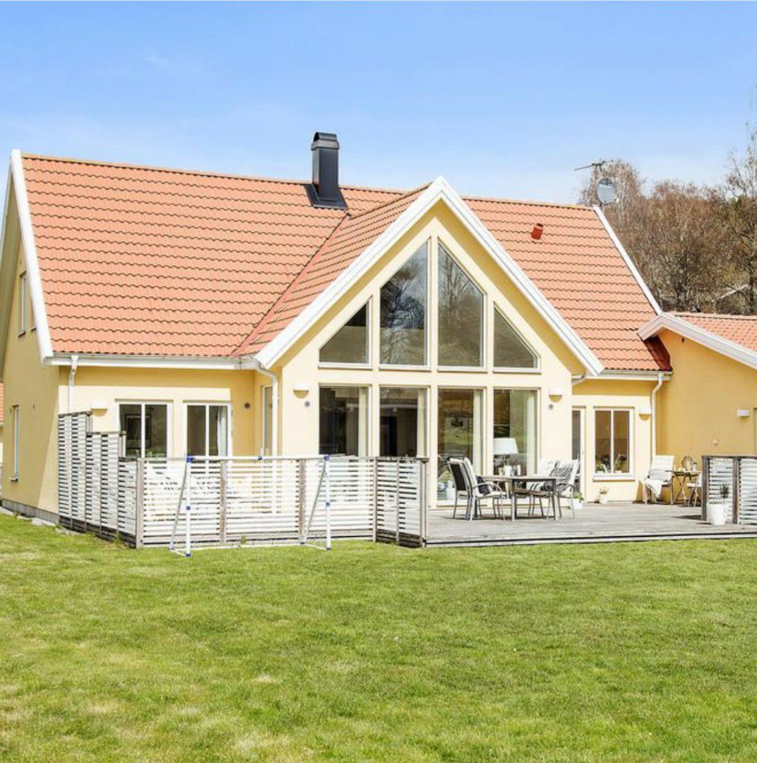A spacious yellow house featuring high ceilings and large triangular windows, allowing ample natural light. The deck is furnished with a dining table and chairs, set against a backdrop of a well-maintained lawn, creating a relaxing outdoor space.