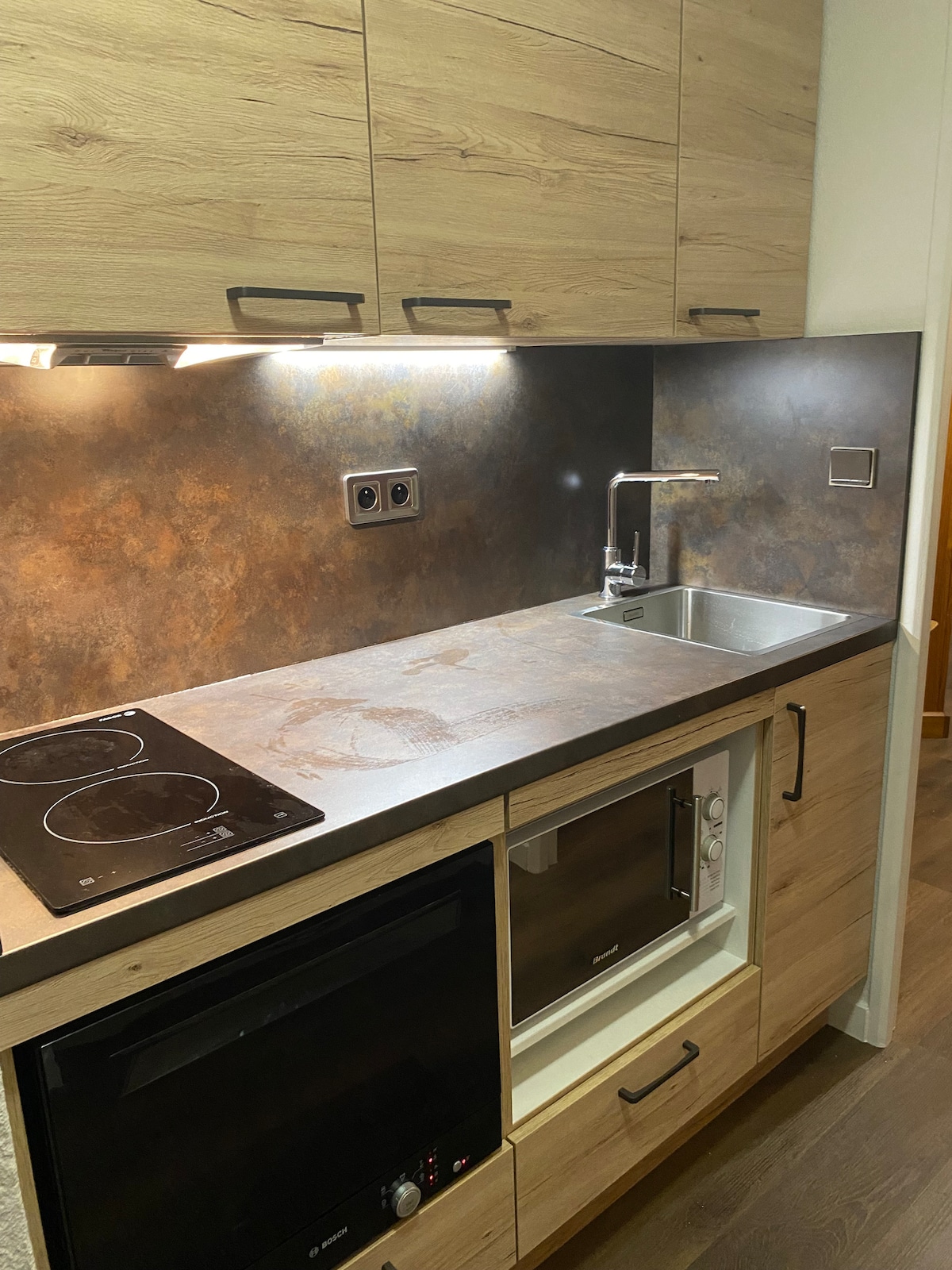 A well-equipped kitchen is featured, showcasing modern appliances including an electric cooktop, microwave, and sink. The cabinetry is finished in a warm wood tone, with ample counter space and soft under-cabinet lighting enhancing visibility.