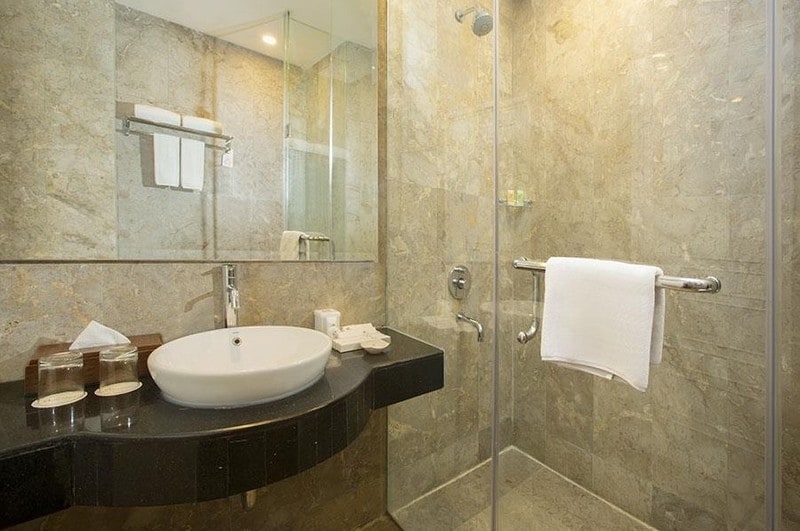 A modern bathroom is presented with elegant stone walls and a glass shower. A minimalist sink is positioned against a dark countertop, featuring toiletries and clean towels. Natural light illuminates the space, enhancing its contemporary design.