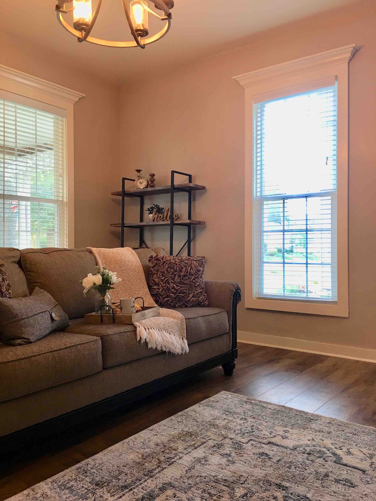 A comfortable seating area is highlighted by a brown sofa adorned with decorative pillows and a soft throw. Natural light filters through a window, illuminating a stylish shelf displaying curated decor. A patterned area rug adds an inviting touch to the wood floor.