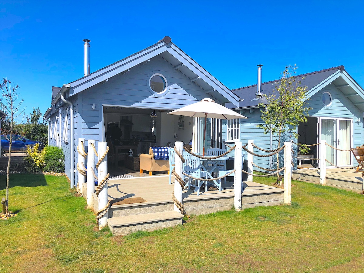 A light blue beach house is featured, set against a clear blue sky. A spacious deck is adorned with a white umbrella, while a comfortable seating area invites relaxation. Lush green grass surrounds the property, contributing to a cheerful outdoor setting.