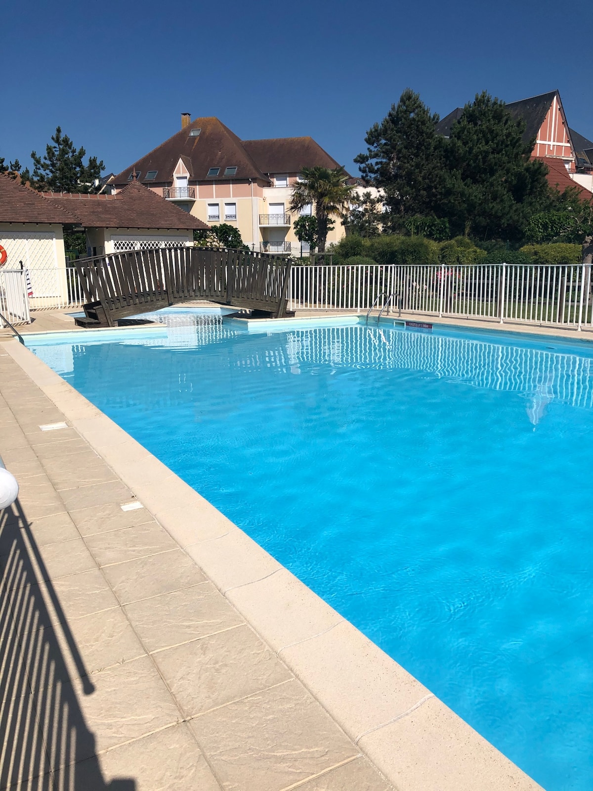 An outdoor swimming pool is surrounded by a secure railing, featuring a clear blue water surface reflecting the sky. Sunlight bathes the area, enhancing the inviting atmosphere. The pool's edge includes steps for easy access, providing a spacious area for relaxation.
