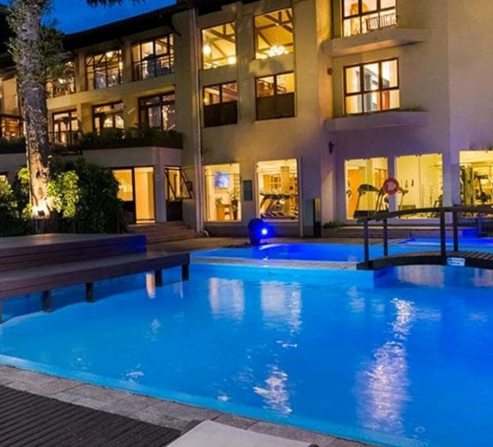 An inviting view of the resort's outdoor pool area is presented, featuring clear blue water and surrounding wooden decks. The building's large windows display illuminated indoor spaces, while trees line the area, adding greenery to the serene environment.