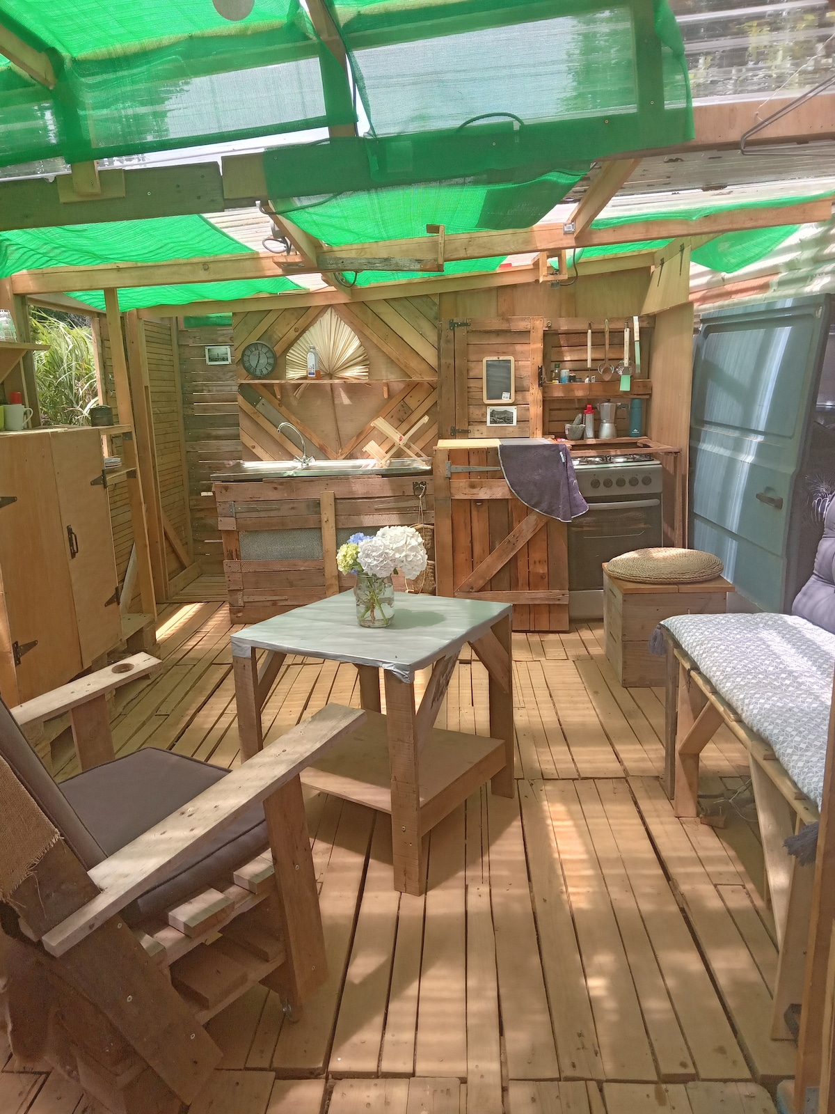 An open kitchen area features a rustic wooden table with a small vase of flowers on top. Surrounding furniture includes a couple of chairs made from repurposed materials. A gas stove is present alongside a wooden countertop, illuminated by natural light filtering through a green canopy.