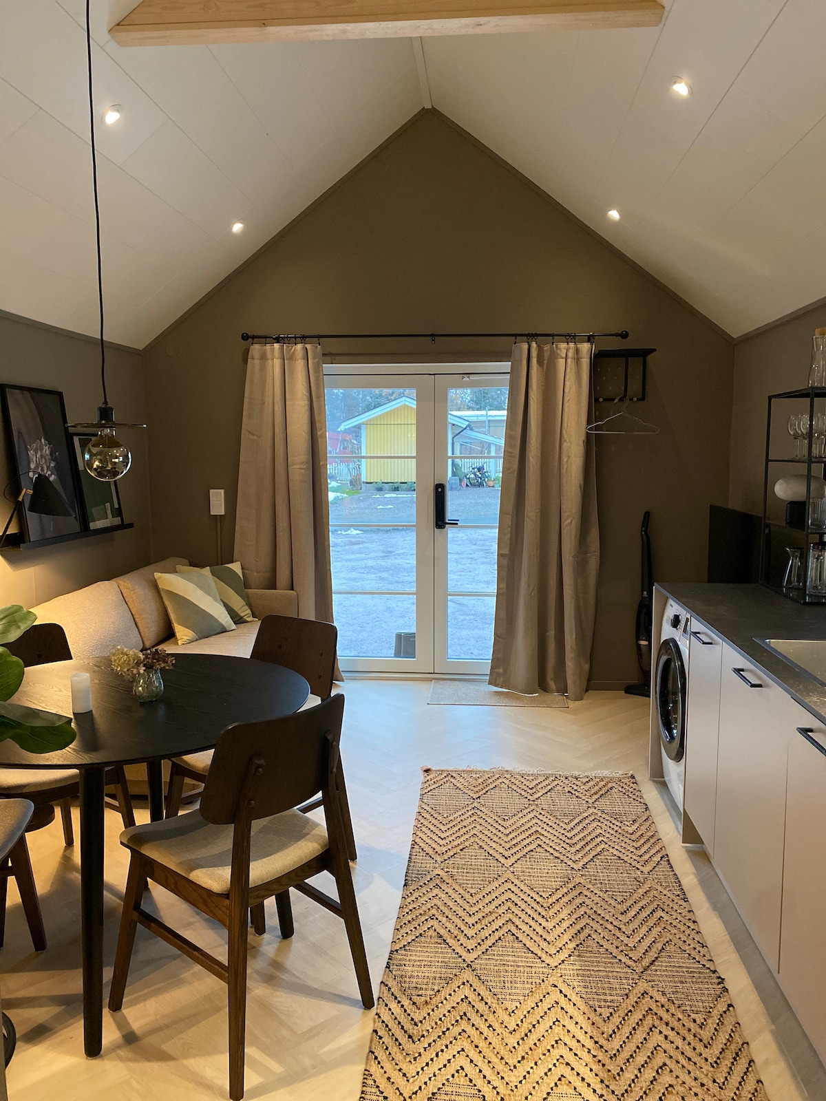 A compact living area features a dining table with four chairs and a sofa against a wall. Large glass doors provide access to the outdoor space, complemented by soft curtains. A light-patterned rug is placed on the floor, and a fully equipped kitchen is visible.