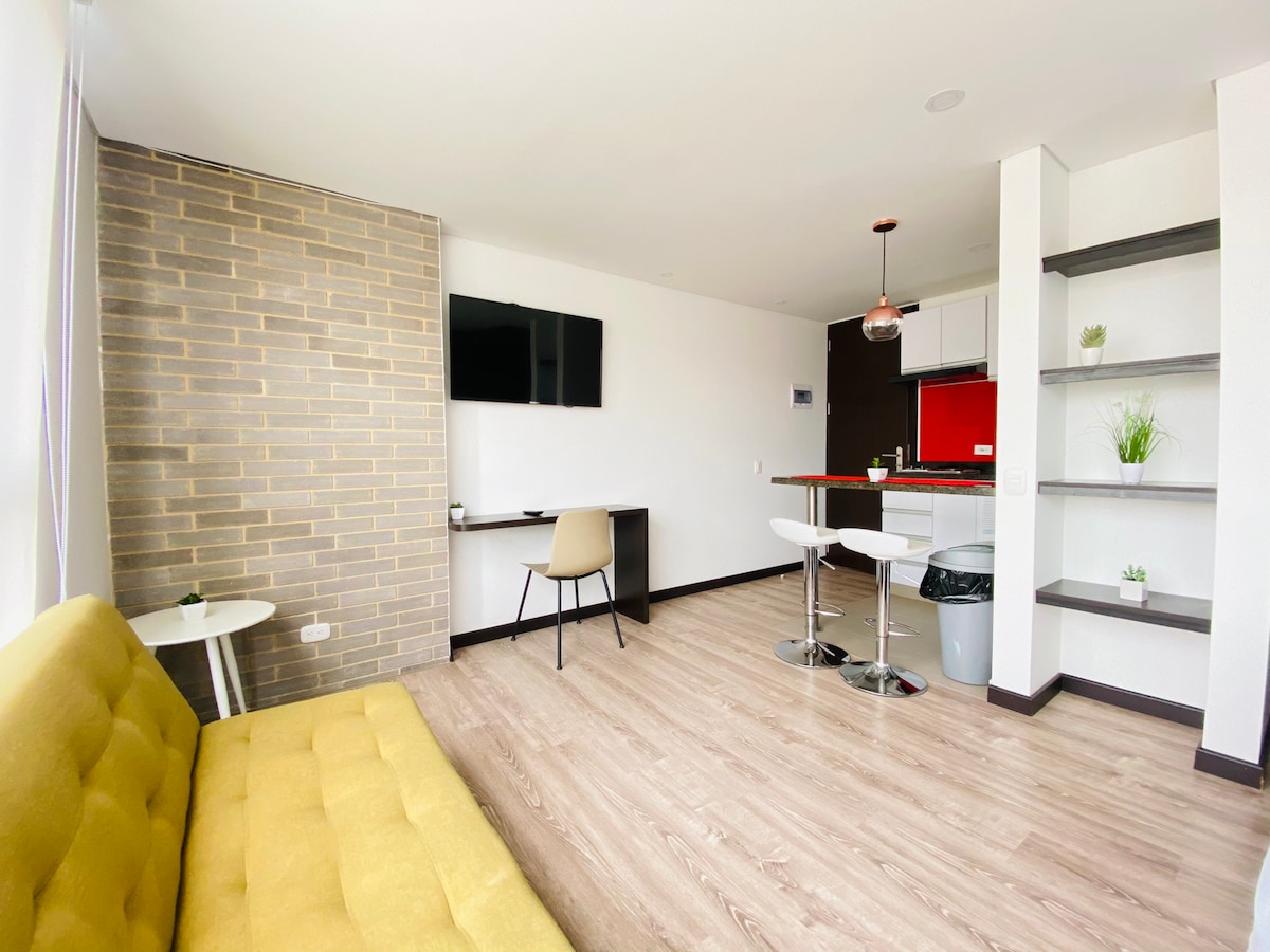 A bright and airy living space features a yellow sofa, a wall-mounted television, and a compact dining area with two stools. The kitchen area is visible, showcasing modern appliances and a red accent wall. Large windows allow natural light to fill the room.