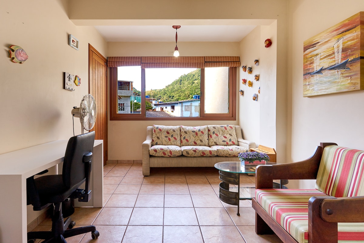 A bright living area features a comfortable floral couch and a striped chair, complemented by a glass coffee table. Natural light pours in through a large window, which offers a view of the surrounding hills. A desk and fan are also situated in this welcoming space.