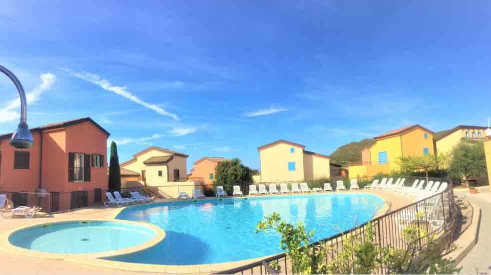A large pool is surrounded by lounge chairs, set against a backdrop of colorful buildings. The calm water reflects the blue sky, while vibrant greenery adds a touch of nature. The area is well-maintained, creating an inviting space for relaxation.