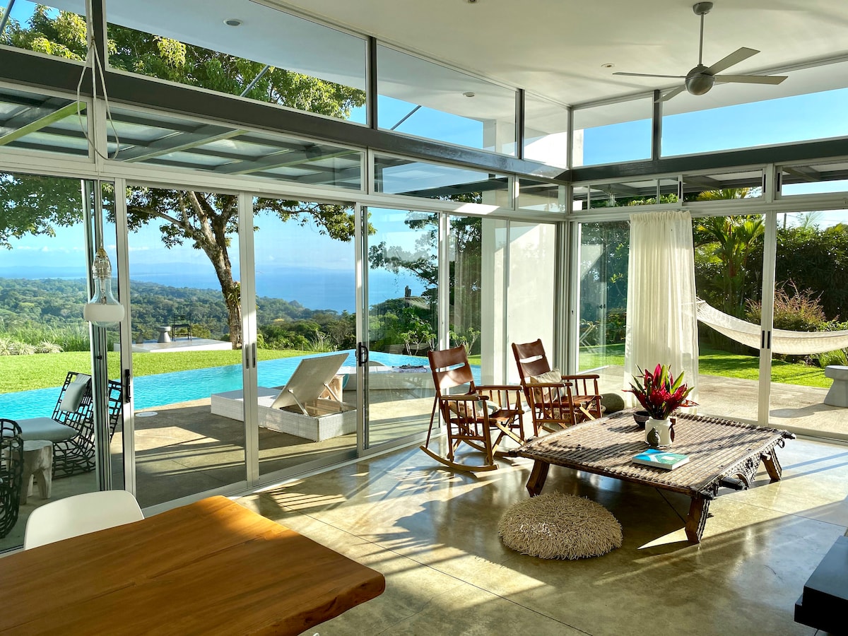 A spacious living area features large glass walls that invite natural light and stunning views of the ocean and landscape. A beautifully arranged seating area includes wooden rocking chairs and a low coffee table, complemented by a dining space with a wooden table. The inviting pool area is visible beyond.