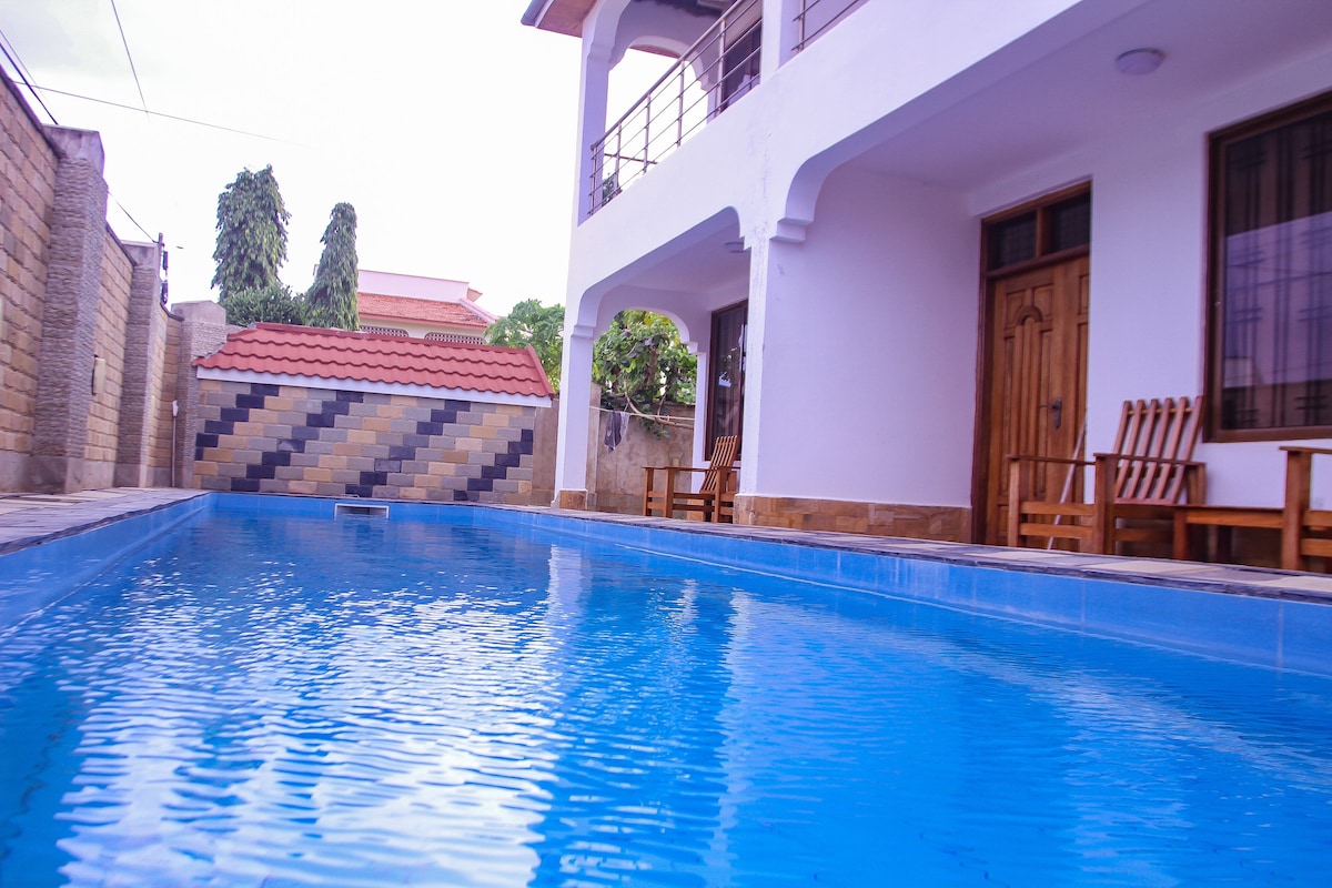 A well-maintained pool is featured, surrounded by a clean, tiled area. Wooden seating is arranged nearby under an overhang, providing a shaded spot for relaxation. The white exterior of the house contrasts with the blue water, creating a calm and inviting environment.