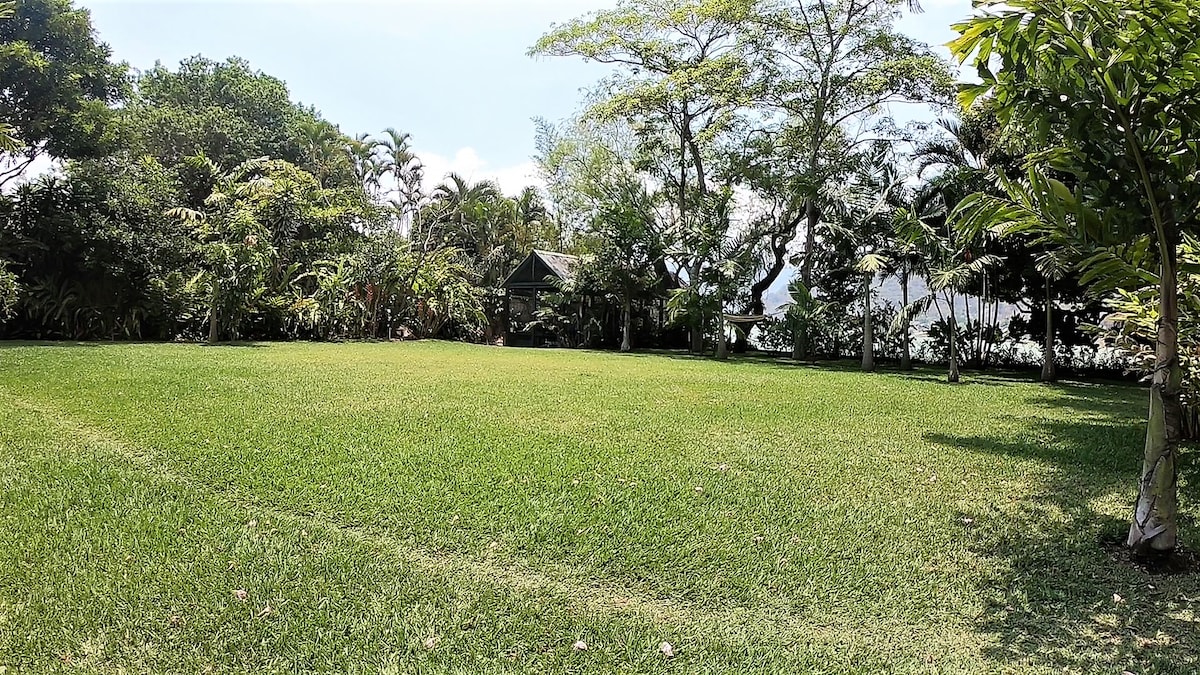 A vast, well-maintained lawn is bordered by lush tropical plants and trees, creating an inviting outdoor space. A glimpse of a small structure is visible in the background. The serene environment offers tranquil views of the surrounding nature.