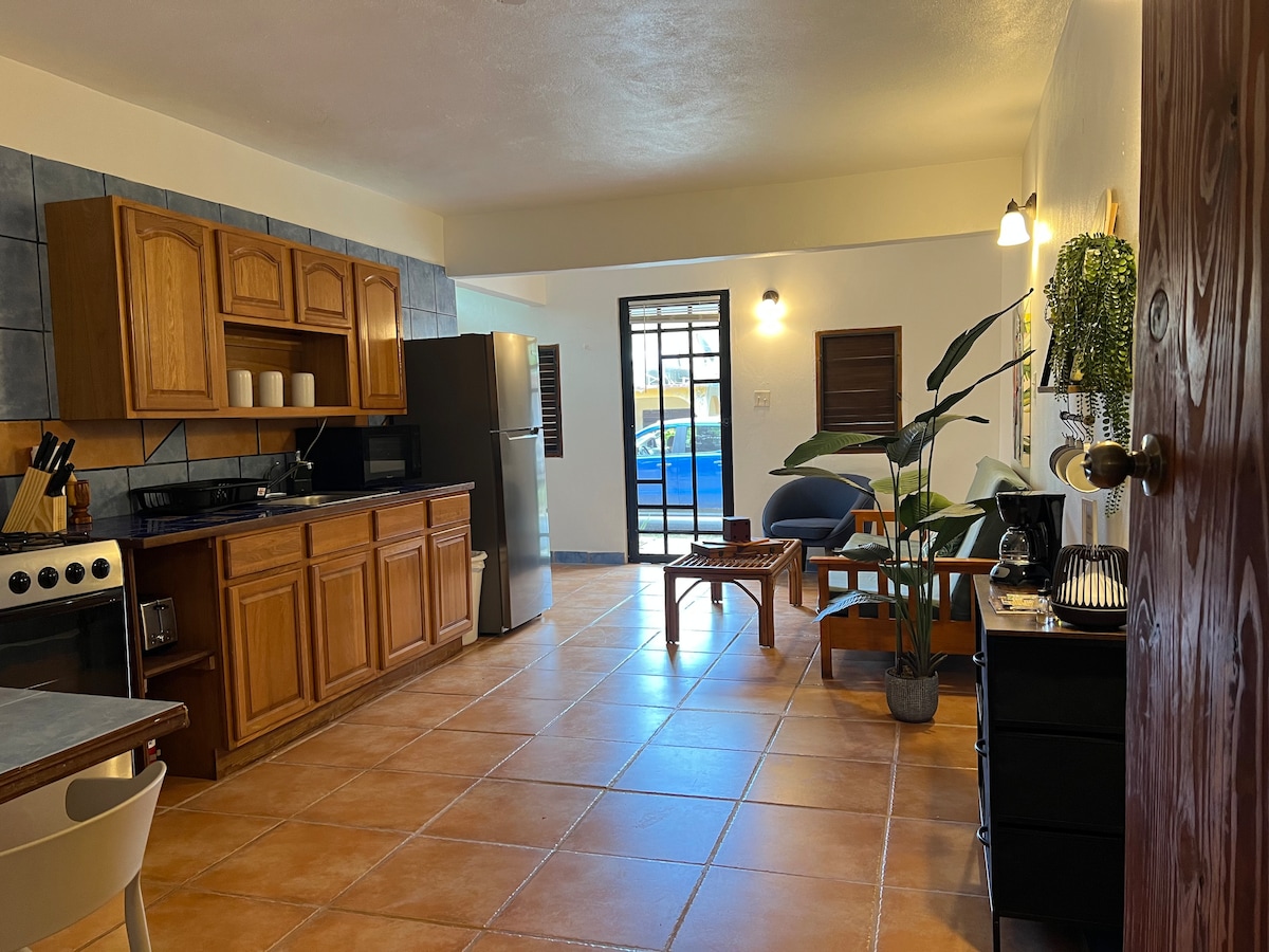 An open-plan kitchen and living area is displayed, featuring wooden cabinetry and tiled floors. A stainless steel refrigerator stands beside a stove. A seating area includes a blue chair and a side table. Natural light enters through a window, illuminating the space.