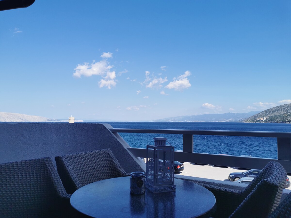 A balcony view showcases a vast blue sea under a clear sky, with distant mountains on the horizon. A round table with a decorative lantern sits beside two woven chairs, inviting relaxation while enjoying the scenic surroundings.