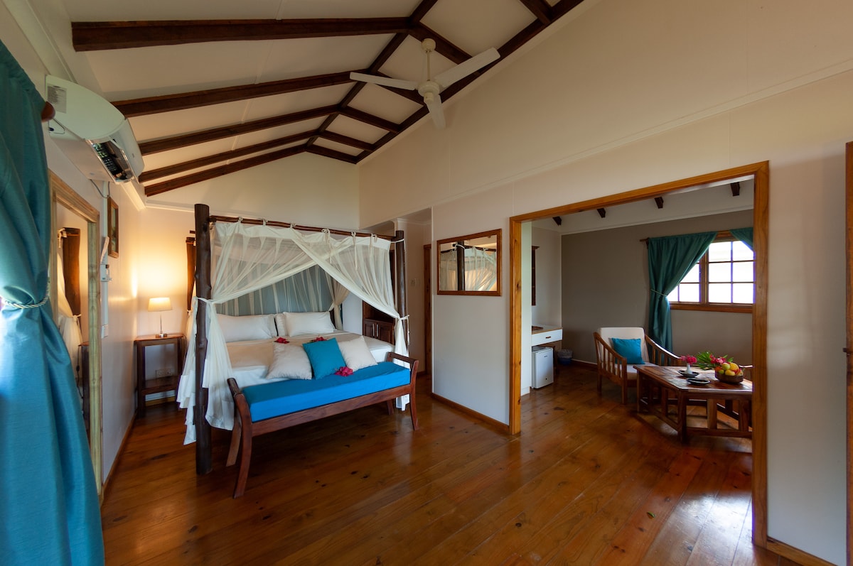 A bright interior features a spacious sleeping area with a wooden canopied bed adorned with white drapery. A seating area is visible through an open doorway, complemented by natural wood accents and soft blue cushions. Large windows allow natural light to fill the room.