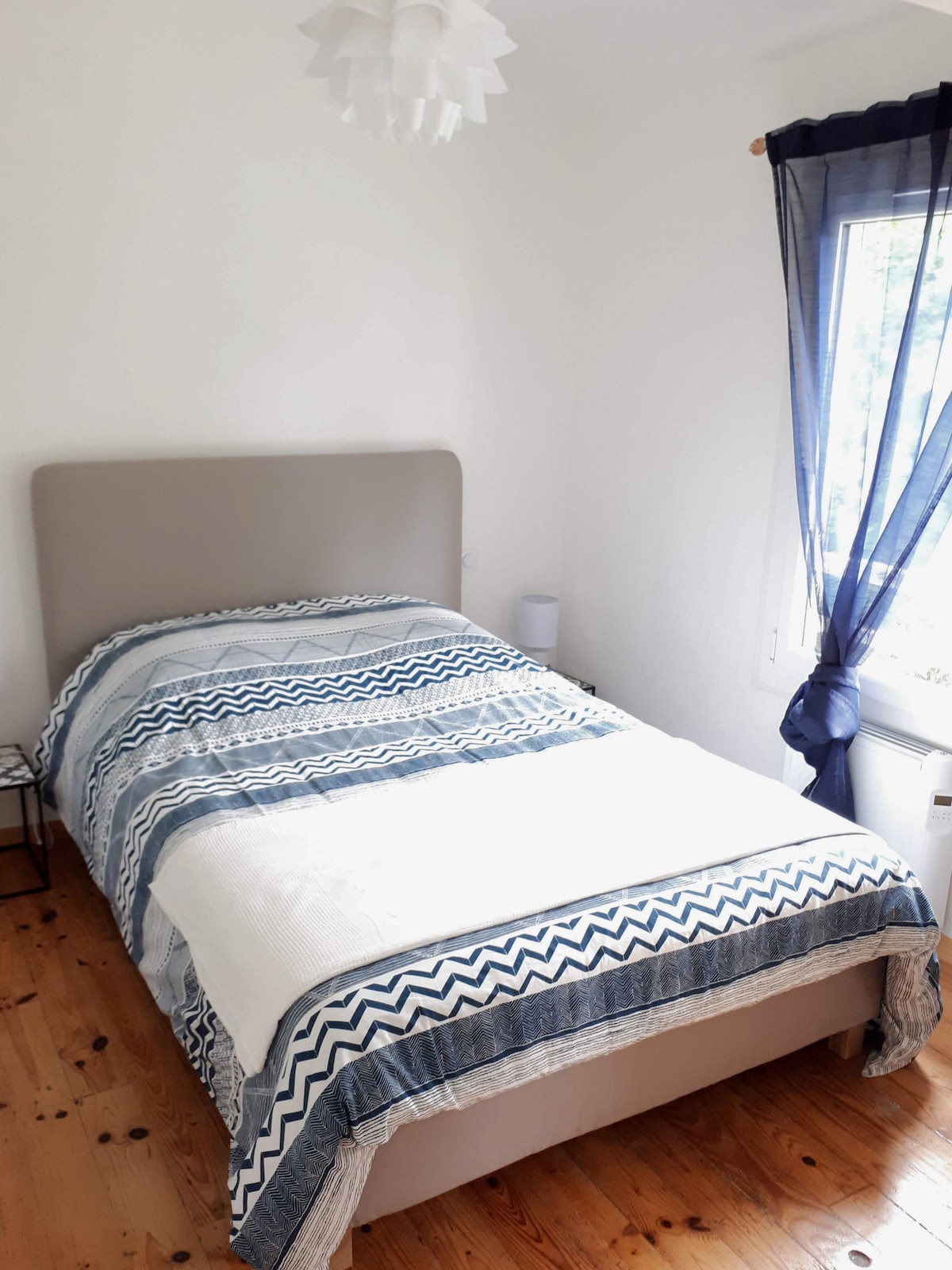 A light-filled bedroom features a spacious bed dressed in a patterned blue and white duvet cover. A simple bedside table is placed alongside, and a window adorned with flowing navy curtains allows natural light to brighten the room.
