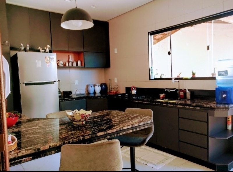 A modern kitchen is presented with dark cabinetry and a large granite countertop. A dining table with four chairs is situated near a window, allowing natural light to illuminate the space. Cooking appliances, a refrigerator, and decorative items enhance functionality.