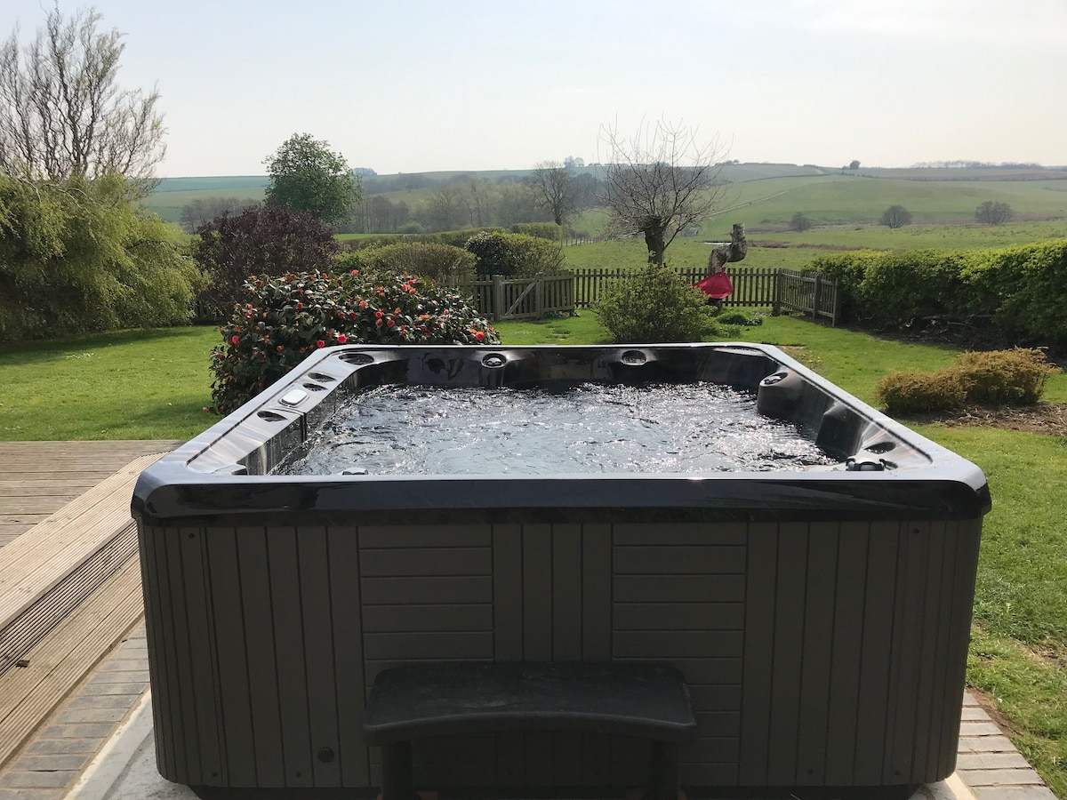 A spacious hot tub is positioned on a wooden deck, offering panoramic views of the rolling landscape. Lush greenery and flowering shrubs surround the area, creating an inviting outdoor space. The background reveals undulating hills, contributing to a serene atmosphere.