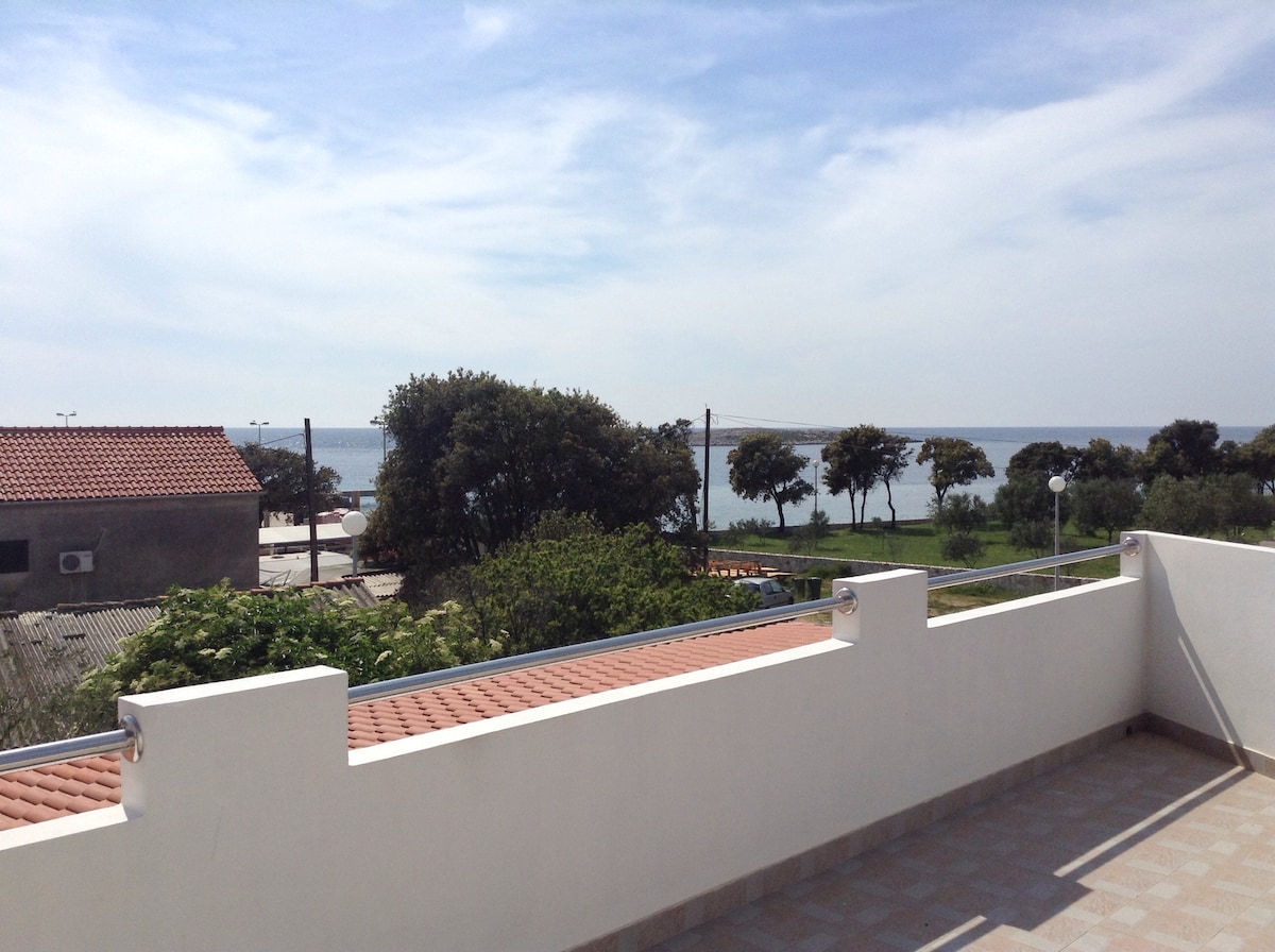 A spacious terrace offers views of the sea and surrounding greenery. The horizon is visible, with trees lining the waterfront. The terrace features a tiled floor, providing a comfortable outdoor setting for relaxation or social gatherings.