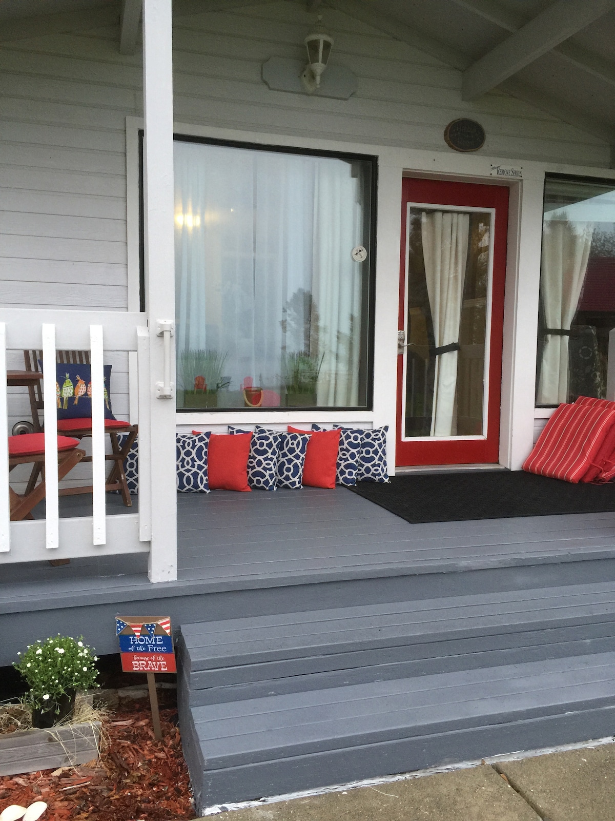 A welcoming porch features comfortable seating arrangements, accented with decorative pillows in blue and red patterns. Light-colored curtains frame the large windows, and a vibrant red door adds a pop of color. Potted plants are visible, enhancing the inviting entryway.