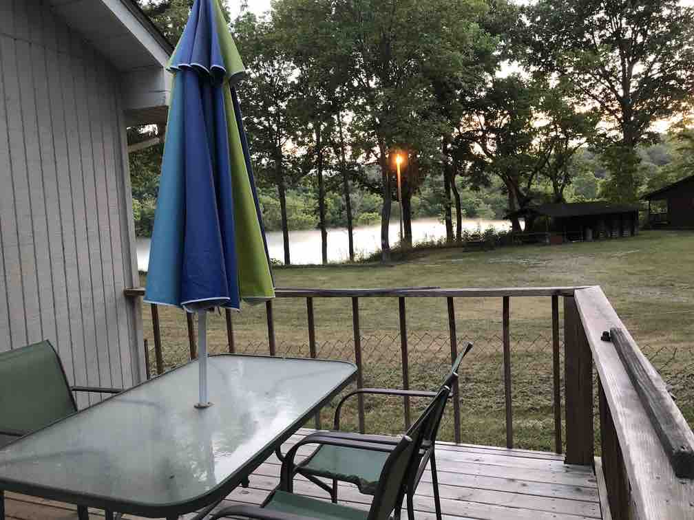A deck is visible featuring a glass top table and chairs, accompanied by a colorful umbrella. The grassy area leads down to the river, surrounded by trees. A soft glow from a nearby light illuminates the serene landscape at dusk.