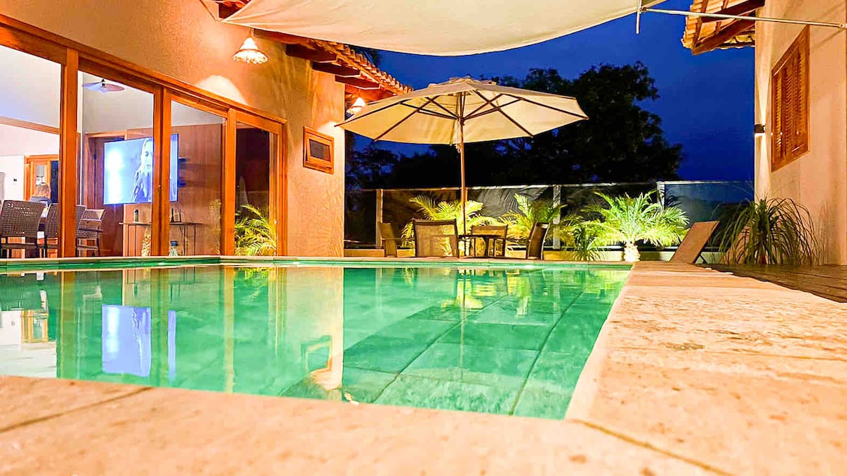 The pool, illuminated in soft lighting, is surrounded by a natural stone deck. An umbrella stands beside a table and chairs set in a cozy corner. Full glass doors lead to the interior, enhancing the connection between outdoor and indoor spaces.