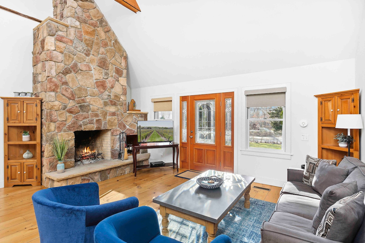 A living room is featured with a stone fireplace as the focal point, surrounded by wooden cabinetry. Large windows invite natural light, and a mix of seating options includes a gray sofa and two blue chairs. A coffee table sits at the center, adorned with a decorative bowl.