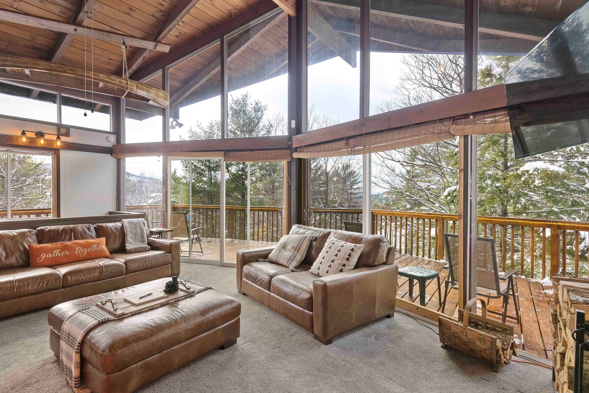 A spacious living area features large windows that invite natural light and provide scenic views of the surrounding nature. Two comfortable sofas are arranged around a central coffee table, with a cozy atmosphere enhanced by the wooden beams of the vaulted ceiling.