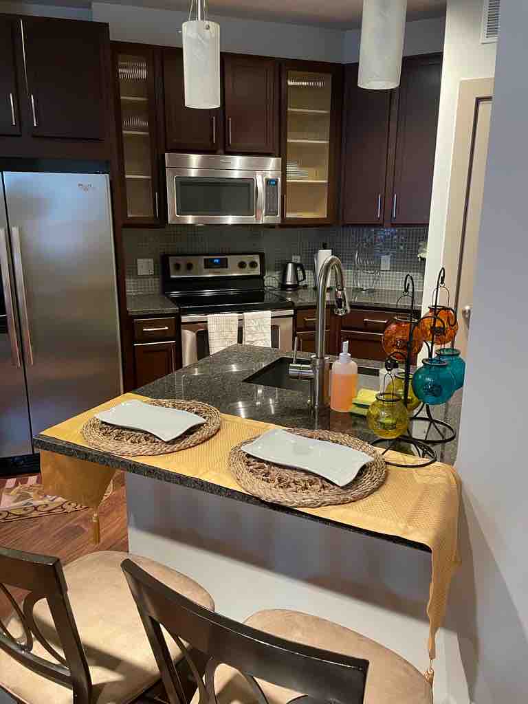A modern kitchen features dark wood cabinetry and stainless steel appliances. A granite countertop is complemented by two place settings, including woven placemats and white plates. Vibrantly colored glass ornaments are arranged on the counter, adding a pop of color.