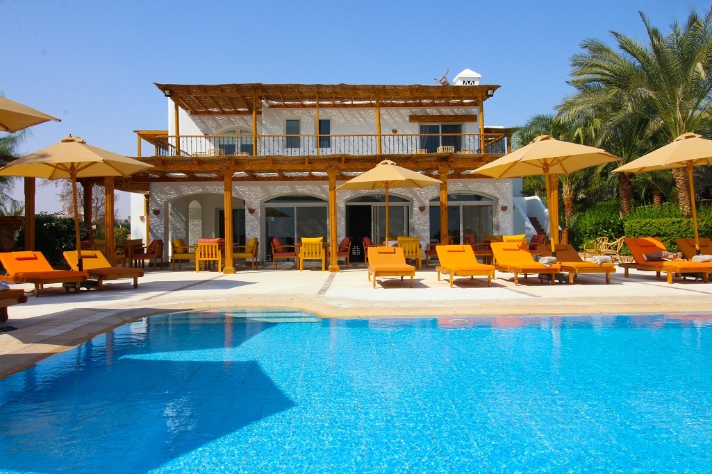 A spacious outdoor area is featured, highlighted by a crystal-clear swimming pool. Surrounding the pool are bright orange lounge chairs arranged under large umbrellas. The building has a wooden pergola and expansive glass doors that lead to the terrace, showcasing a serene atmosphere with palm trees in the background.