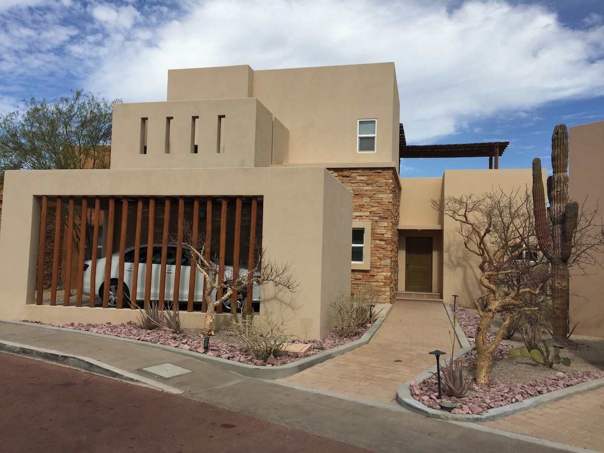 The exterior of the two-floor modern house is highlighted by natural stone and wood elements. A pathway leads to the entrance, flanked by desert landscaping with low shrubs, rocks, and cacti. Features include wooden slats and a large window on the upper floor for ample light.