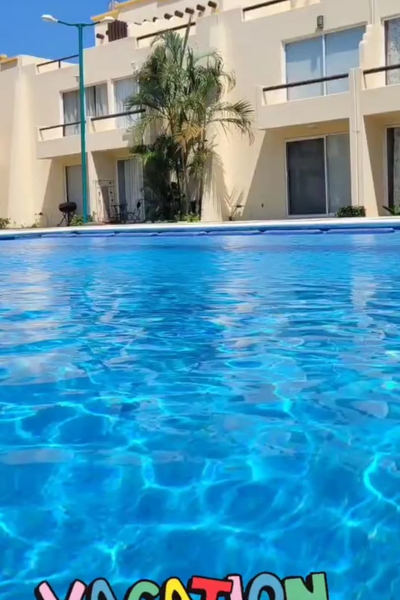 A clear swimming pool is visible in the foreground, with rippling blue water reflecting sunlight. The background features a beige building with large glass doors, surrounded by greenery. The setting provides a sense of direct access to the pool from the ground floor.