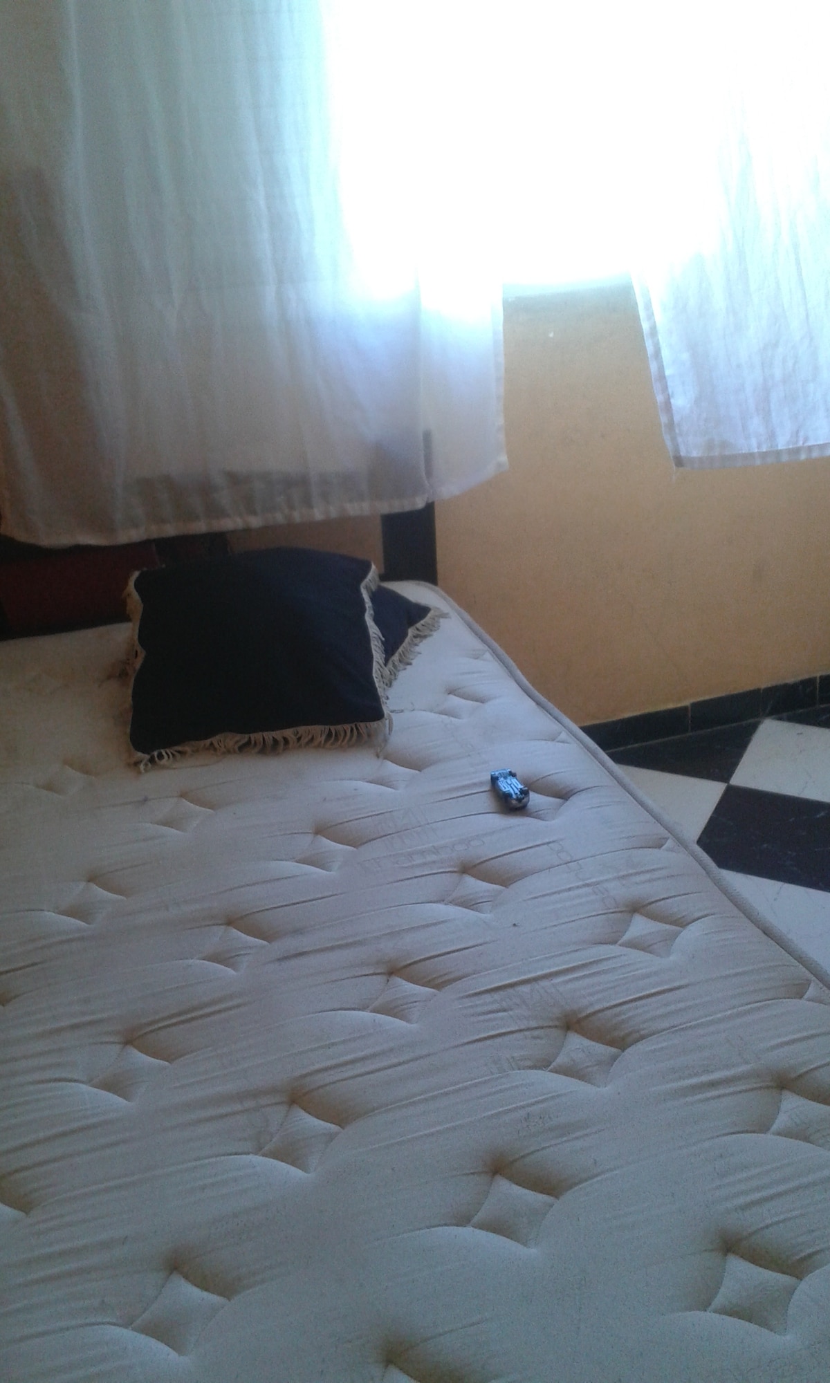 A single bed with a light-colored mattress is positioned against a wall, accompanied by a dark pillow. Natural light filters through sheer curtains, creating a soft ambiance in the room, while a small item rests on the bed's surface.