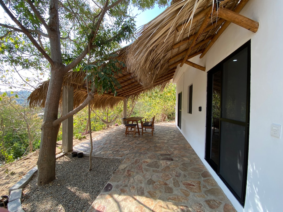 A covered patio area is shown, featuring a wooden table surrounded by chairs. The natural stone floor adds texture, while a thatched roof provides shade. Lush greenery surrounds the space, enhancing the tranquil environment.