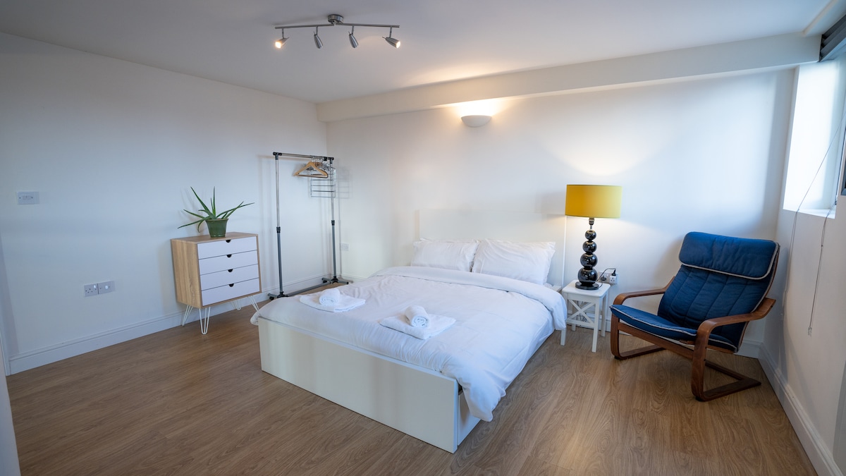 A well-sized bedroom offers a spacious bed neatly made with fresh linens and two folded towels. A bedside lamp illuminates the area, while a clothing rack and dresser provide storage. Natural light is allowed through a large window, enhancing the clean, minimalist aesthetic.
