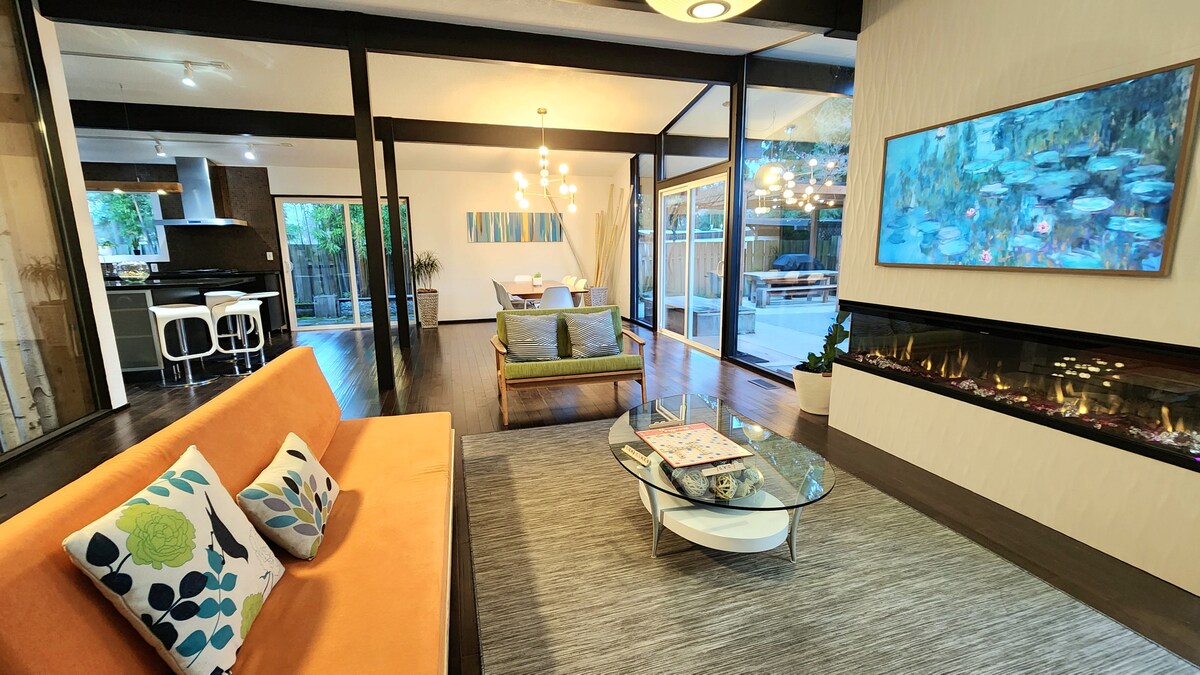 An open living space features a stylish orange sofa adorned with decorative pillows. A modern glass coffee table sits on a plush area rug in front of a wall-mounted artwork. Natural light floods through floor-to-ceiling windows, highlighting the cozy electric fireplace.