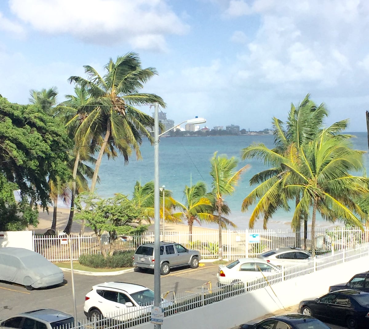 A view of the beach is framed by swaying palm trees, revealing the turquoise ocean in the background. Vehicles are parked along the street, and a white fence separates the road from the sandy shore, providing a clear connection to the coastal landscape.