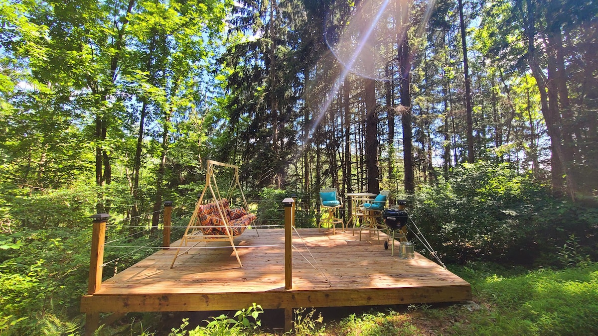 A wooden deck situated among tall trees provides space for relaxation. A cozy swing chair is positioned on one side, while a small table and two blue chairs invite guests to enjoy outdoor meals. Natural light filters through the surrounding greenery.