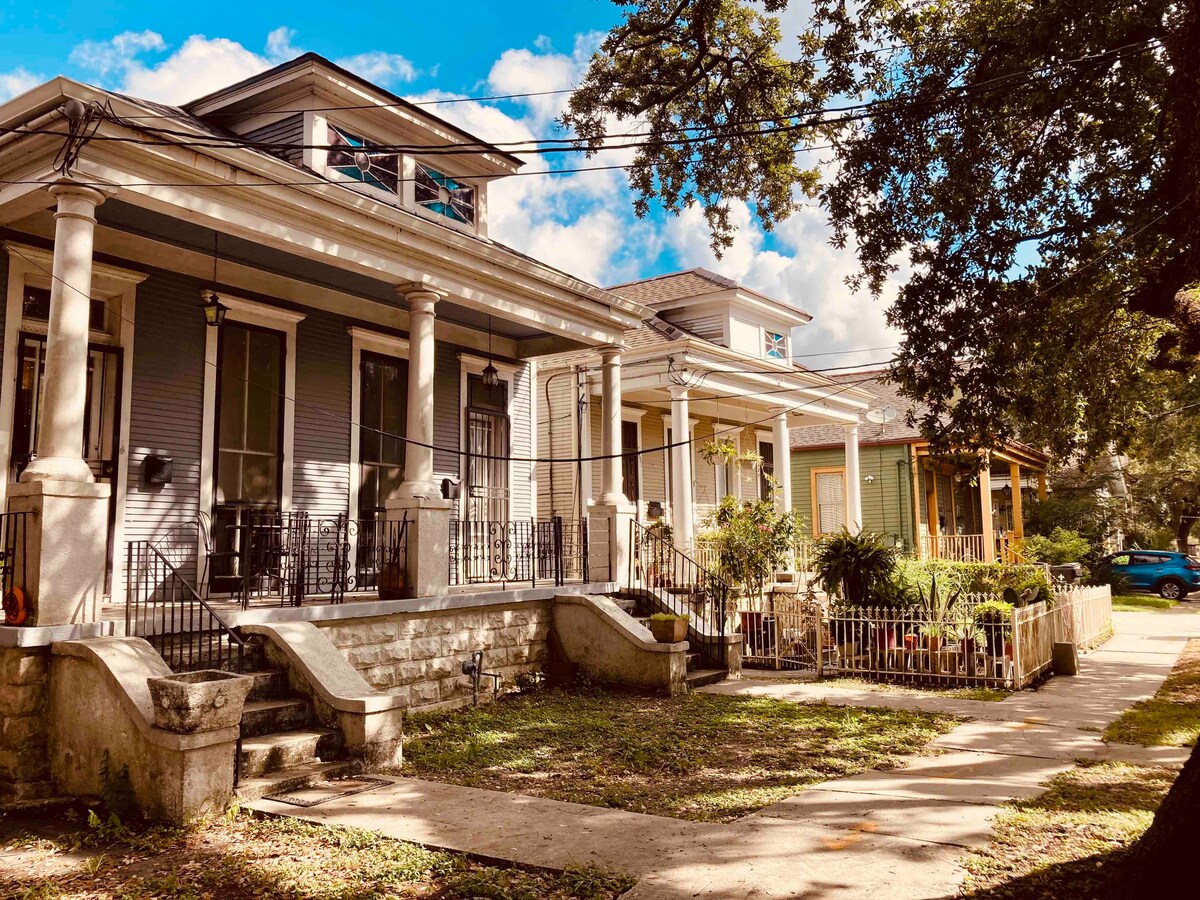 A charming exterior view of the property features classic columns and a spacious porch. Lush greenery surrounds the walkways, with sunlight filtering through the trees. The architecture reflects the character of the neighborhood, inviting exploration of the vibrant surroundings.