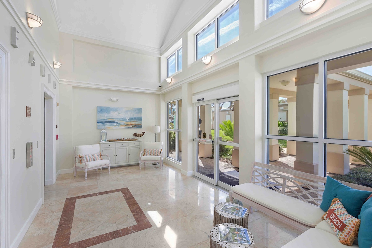 A bright lobby area features tall windows allowing natural light to fill the space. Elegant light fixtures are mounted on the walls. Comfortable seating options, including two chairs and a bench, are arranged around a decorative coffee table. A beach-themed painting adds a serene touch.