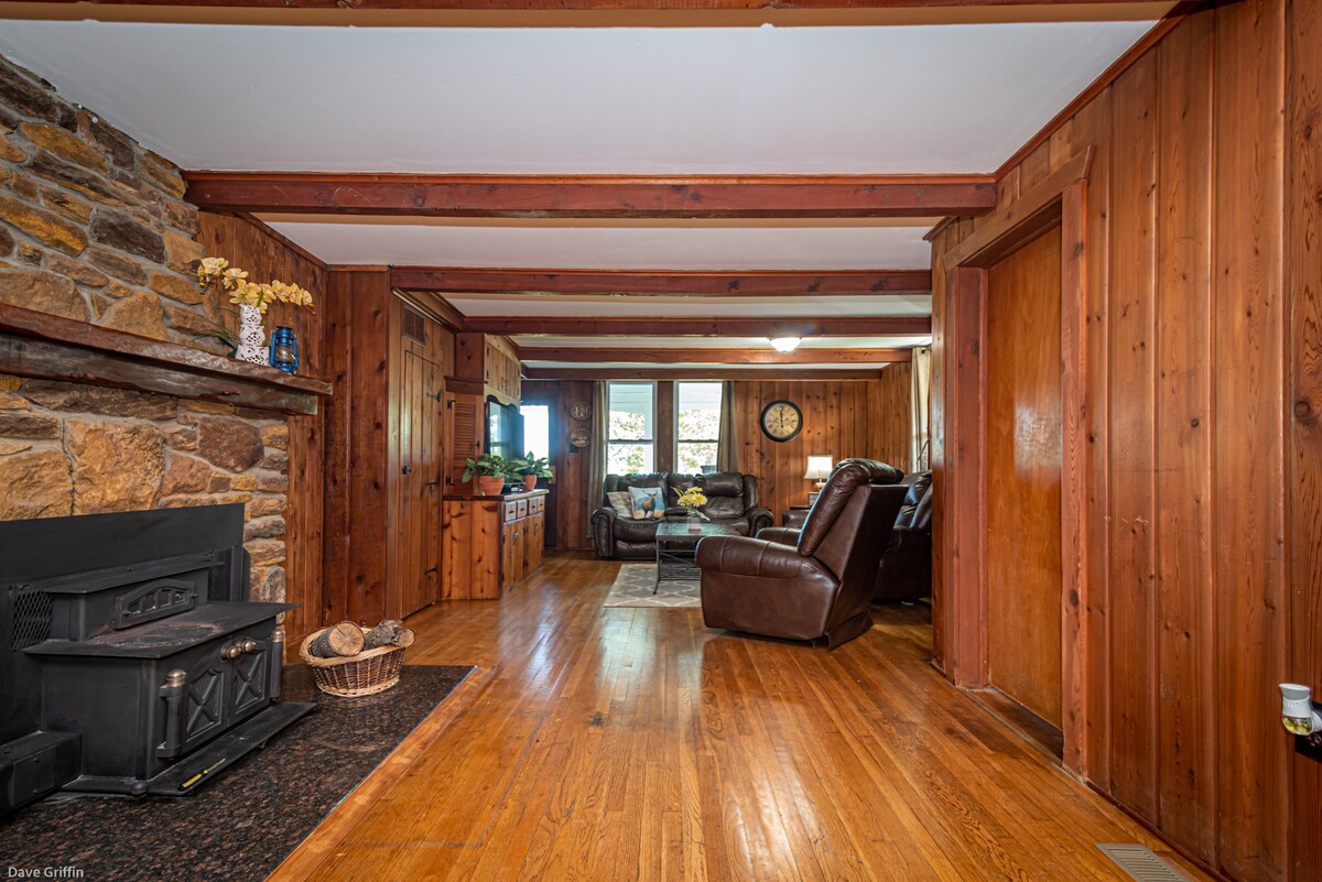 A wooden interior features exposed beams and a stone fireplace, creating a rustic charm. Dark leather seating is arranged in a cozy living area, while large windows allow natural light to fill the space. Warm wood floors enhance the inviting atmosphere.