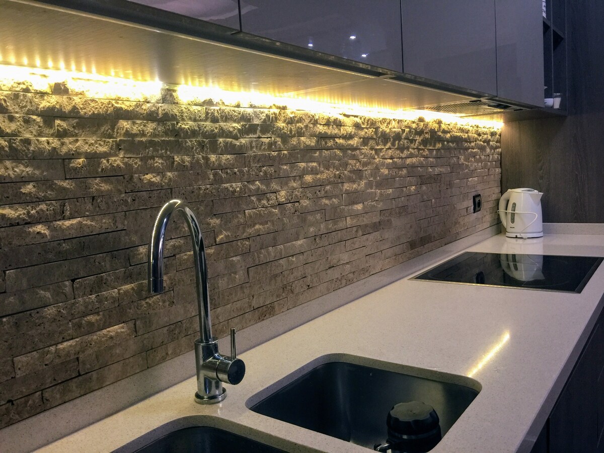 A sleek kitchen space features a polished countertop with two integrated sinks. A textured stone wall is illuminated by warm under-cabinet lighting. Modern appliances, including a kettle, are positioned nearby, enhancing the kitchen's contemporary aesthetic.